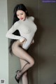 A woman in a white bodysuit and black stockings leaning against a wall.