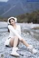 A woman sitting on a rocky beach wearing a white sweater and ear muffs.